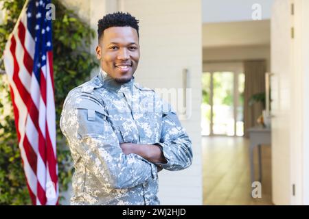 Männlicher afroamerikanischer Soldat, der mit Armen vor dem Haus steht, mit us-Flagge, Kopierraum Stockfoto