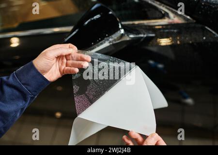 Zugeschnittene Ansicht eines engagierten Spezialisten in bequemer blauer Uniform, der Schutzfolie auf dem Spiegel des Autos aufträgt Stockfoto