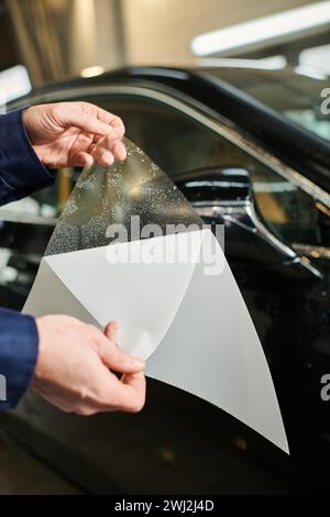 Zugeschnittene Ansicht eines hart arbeitenden Spezialisten in blauer Uniform, bereit zum Auftragen von Schutzfolie auf Autospiegel Stockfoto