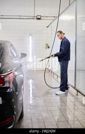 Attraktiver Berufskollegen in blauer Uniform, der in der Garage schwarzes Auto mit Schlauch wäscht Stockfoto