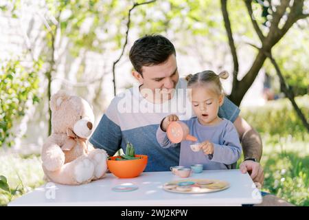 Das kleine Mädchen gießt Tee aus einer Spielzeug-Teekanne in einen Spielzeug-Becher, während es auf dem Schoß ihres Vaters am Tisch sitzt Stockfoto