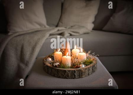 Vier weiße Adventskerzen und Weihnachtsdekoration in einem rustikalen Baumtablett auf einer Ottomane auf einer Couch, gemütliche Winterdekoration im Stockfoto