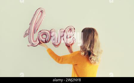 Glückliche junge Frau, die glänzendes rosa Wort Liebe hält, Buchstaben geformte Folienballons auf weißem Wandhintergrund Stockfoto