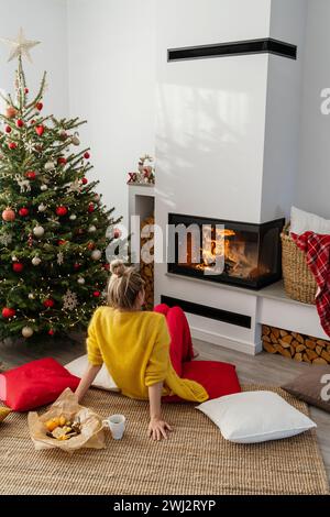 Glückliche Frau sitzt neben einem glühenden Kamin in einem gemütlichen Wohnzimmer, geschmückt mit einem Weihnachtsbaum Stockfoto