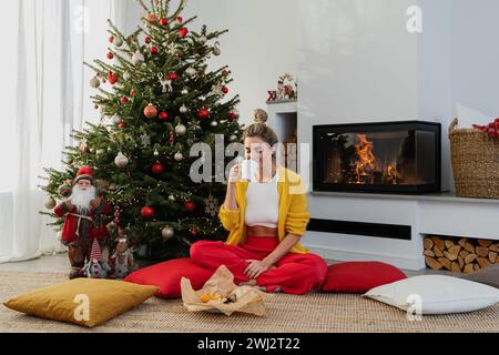 Glückliche Frau sitzt neben einem glühenden Kamin in einem gemütlichen Wohnzimmer, geschmückt mit einem Weihnachtsbaum Stockfoto