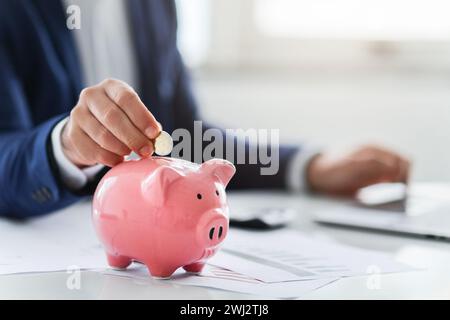 Fokussiertes Bild einer Hand eines Geschäftsmannes, die eine Münze in eine rosa Sparkasse einlegt Stockfoto