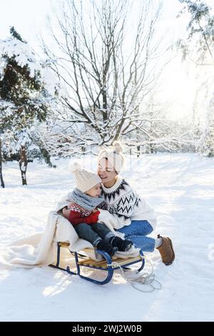 Junge Mutter und ihr süßer kleiner Sohn mit Retro-Schlitten in einem verschneiten Park an sonnigen Tagen Stockfoto