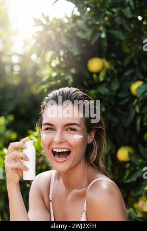 Fröhliche und schöne Frau, die feuchtigkeitsspendende Creme oder Sonnencreme auf ihre Gesichtshaut aufträgt Stockfoto