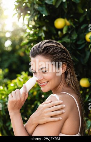 Fröhliche und schöne Frau, die Feuchtigkeitscreme oder Sonnencreme auf ihren Körper aufträgt Stockfoto