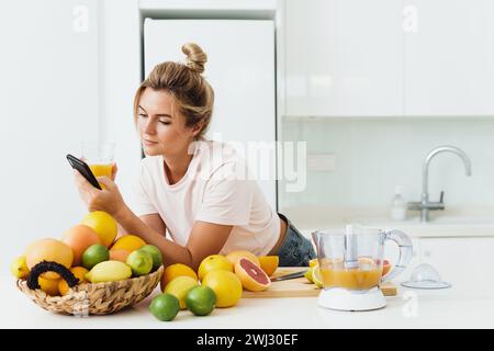 Junge Frau, die frisch gepressten, selbstgemachten Orangensaft trinkt und ihr Smartphone benutzt Stockfoto