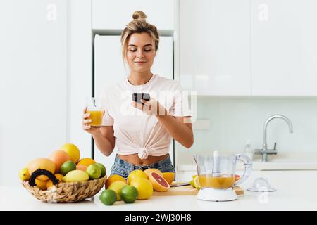 Frau, die frisch gepressten, selbstgemachten Orangensaft trinkt und Fotos mit ihrem Smartphone macht Stockfoto