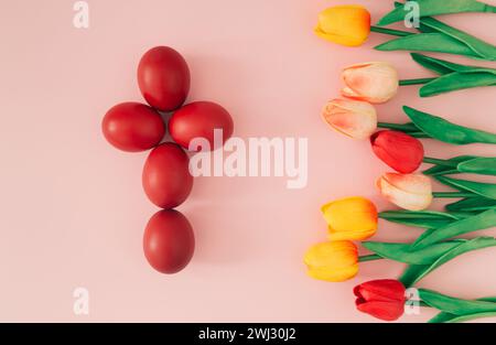 Kreative Osterfestkomposition aus roten Ostereiern in Form eines Kreuzes und bunten Tulpen auf pastellrosa Hintergrund. Minimales Osterkonzept. Stockfoto