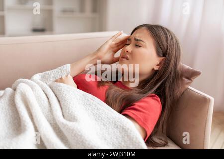 Unbequeme junge asiatische Frau, die auf der Couch liegt und den Tempel berührt Stockfoto
