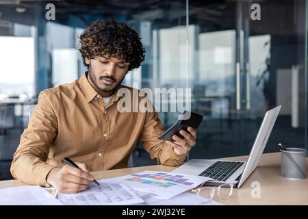 Ernsthafter und zielgerichteter junger Indianer, der im Büro mit Dokumenten arbeitet. Verarbeitet Daten und Konten, verwendet ein Notizbuch und einen Taschenrechner. Stockfoto