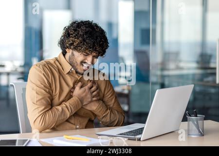 Herzinfarkt und Panikattacke am Arbeitsplatz. Junger indischer Mann mit Brustschmerzen, der am Schreibtisch im Büro sitzt und die Hände hält. Stockfoto