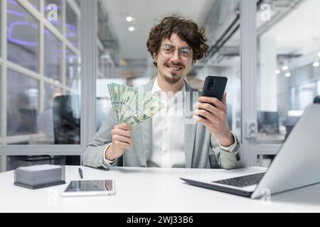 Porträt eines glücklichen jungen Geschäftsmannes, der am Schreibtisch im Büro sitzt, Telefon und Geld hält und zeigt, lächelt und in die Kamera schaut. Stockfoto