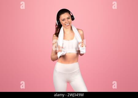 Eine lächelnde, athletische Frau, die einen weißen Sport-BH und Leggings mit Kopfhörern trägt und ein Handtuch über die Schultern hält Stockfoto