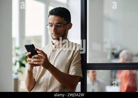 Junger muslimischer Büromanager oder Ökonom, der sich den Smartphone-Bildschirm anschaut, während er im Coworking-Raum gegen kommunizierende Kollegen steht Stockfoto