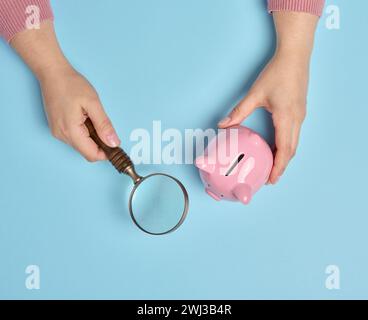 Die Hand einer Frau hält eine Schweinebank und eine Lupe auf blauem Hintergrund Stockfoto