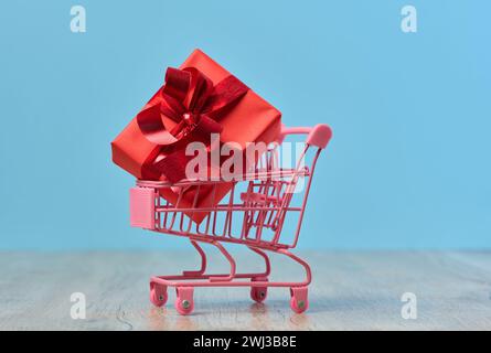 Ein in rotes Papier gewickeltes Geschenk in einem Miniatur-Einkaufswagen auf blauem Hintergrund Stockfoto