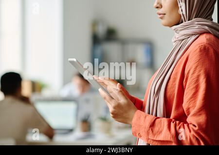 Zugeschnittene Aufnahme einer jungen muslimischen Managerin, die ein Tablet benutzt, während sie im Coworking-Bereich gegen männliche Kollegen steht Stockfoto