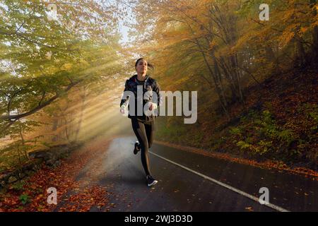 Marathonläuferin trainiert an einem kalten Herbsttag auf einer Straße durch einen nebeligen Wald mit Sonnenstrahlen Stockfoto