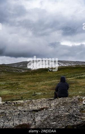 Einsames Kind, das auf felsigem Felsvorsprung sitzt und die weite zerklüftete Landschaft und den weiten Blick auf die weiße Klippe am bewölkten Himmel in Norwegen betrachtet Stockfoto