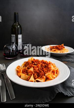 Schüsseln Pappardelle Nudeln in authentischer Bolognese-Sauce: Nahaufnahme flacher Nudelschüsseln mit einem Glas Rotwein, Gabeln und Löffeln Stockfoto