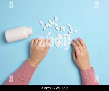 Die Hand einer Frau, die eine Plastikflasche und weiße ovale Pillen auf blauem Hintergrund hält, von oben. Stockfoto