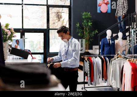 Asiatischer Mitarbeiter im Geschäft arrangiert Kleidung, legt modische Waren auf Kleiderbügel, arbeitet in modernen Boutique Visual. Stilvoller Mann, der formelle Kleidungsstücke im Einkaufszentrum prüft. Modekonzept Stockfoto