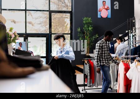 Ladenarbeiter mit stylischen Hemden und modischen Waren auf Kleiderbügeln im Einkaufszentrum. Fröhlicher Manager arrangierte Freizeitkleidung und prüfte den Preis für Kleidung in einer modernen Boutique Stockfoto