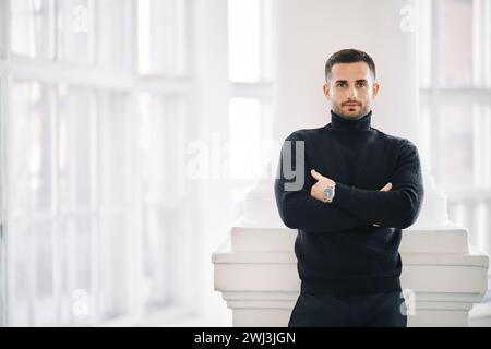 Ein selbstbewusster, gutaussehender Mann in einem schwarzen Rollkragen steht mit überkreuzten Armen im modernen Innenraum Stockfoto