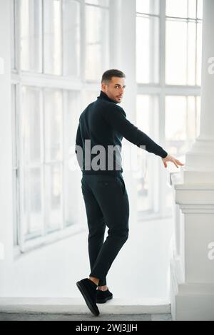 Hübsches modisches Männermodell in einem komplett schwarzen Kleidungsoutfit, das auf einer weißen Treppe posiert Stockfoto