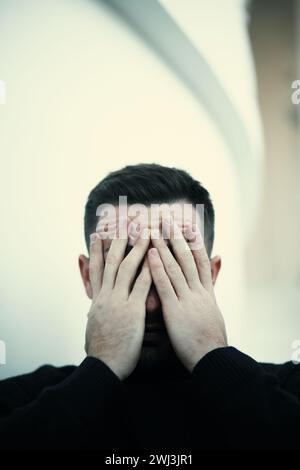 Ein junger Mann mit einem dunklen Rollkragen bedeckt sein Gesicht mit den Händen in einem nachdenklichen oder emotionalen Moment Stockfoto