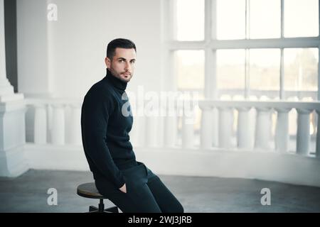 Trendiger, gutaussehender Mann sitzt nachdenklich auf einem Stuhl in einem geräumigen weißen Zimmer Stockfoto