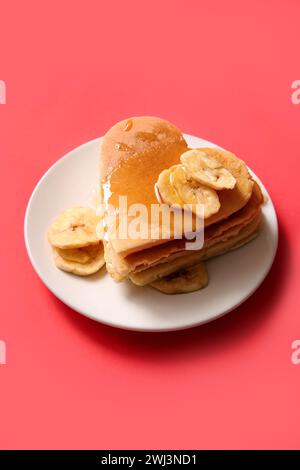 Teller mit leckeren Pfannkuchen in Herzform, getrockneter Banane und Honig auf rotem Hintergrund. Valentinstag-Feier Stockfoto