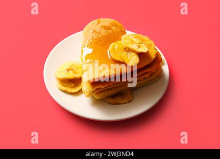 Teller mit leckeren Pfannkuchen in Herzform, getrockneter Banane und Honig auf rotem Hintergrund. Valentinstag-Feier Stockfoto