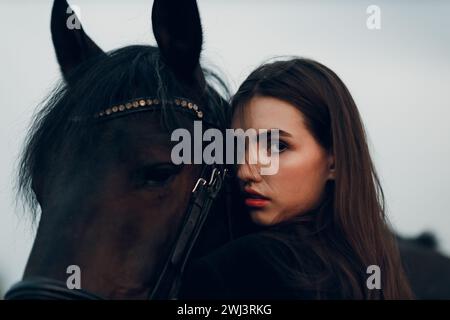 Junge Frau in Schwarz mit ihrem Pferd bei Sonnenuntergang draußen. Stockfoto