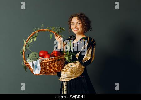 Porträt einer jungen erwachsenen Frau in einem mittelalterlichen Kleid, die einen Korb mit Gemüse und Früchten in der Hand hält. Stockfoto