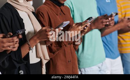Eine vielfältige Gruppe von Schülern, die in das digitale Zeitalter eingetaucht sind, steht vereint, während sie mit ihren Smartphones gegen eine weiße Ba interagieren Stockfoto