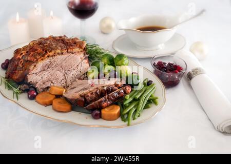 Schweinebraten mit Kruste, grünen Bohnen, Rosenkohl, Karotten, Preiselbeere und Soße auf weißem Tischtuch mit Kerzen und Chri Stockfoto