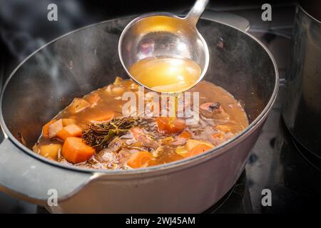 Klare Brühe wird mit einer Kelle in einen Topf mit dampfendem Gemüse wie Karotten, Zwiebeln, Sellerie, Lauch und Kräutern gegossen Stockfoto