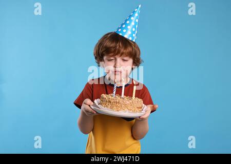 Geburtstagsfeier. Netter kleiner Junge im Partyhut, der Kerzen auf leckerem Kuchen vor hellblauem Hintergrund bläst Stockfoto