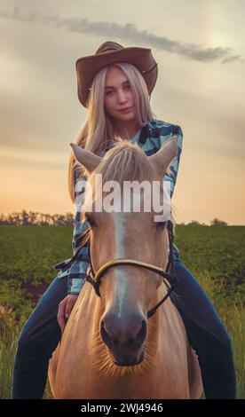 Ein junges Mädchen mit Cowboyhut und blauer Jeans sitzt auf einem Pferd vor dem Hintergrund des Sonnenuntergangs auf der Ranch Stockfoto