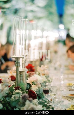 Brennende Kerzen auf Kerzenleuchtern mit Glaskappen stehen auf einem festlichen Tisch zwischen Blumen Stockfoto