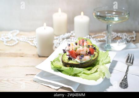 Salat aus Garnelen, Tomaten und Mayonnaise gefüllt in einer halben Avocado auf einem hellen rustikalen Holztisch, Weinglas, weiße Kerzen Stockfoto