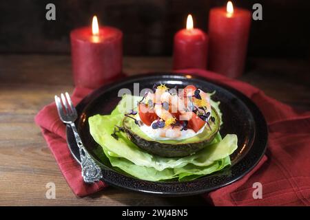 Halbe Avocado gefüllt mit Garnelen, Tomaten und Mayonnaise auf einem schwarzen Teller, rote Kerzen und Serviette, dunkles rustikales Holzhintergras Stockfoto