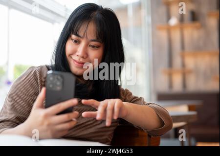 Eine positive asiatische Frau liest SMS auf ihrem Smartphone, während sie sich in einem Café entspannt. Konzepte für Menschen und drahtlose Technologie Stockfoto
