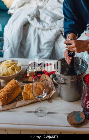 Frau in blauem Hemd, die eine Flasche gekühlten Wein in Sektkübel auf dem Kaffeetisch mit Snacks öffnet Stockfoto