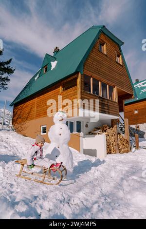 Das kleine Mädchen sitzt auf einem Holzschlitten neben einem Schneemann in der Nähe eines hölzernen Häuschens. Seitenansicht Stockfoto